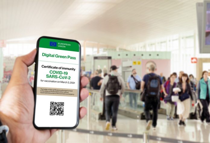 The digital green pass of the european union with the QR code on the screen of a mobile held by a hand with a blurred airport in the background